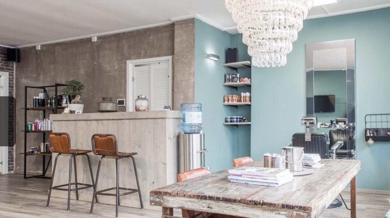 De salon van binnen: de bar met leren krukken, de houten tafel, de kroonluchter en de spiegel met de kappersstoel